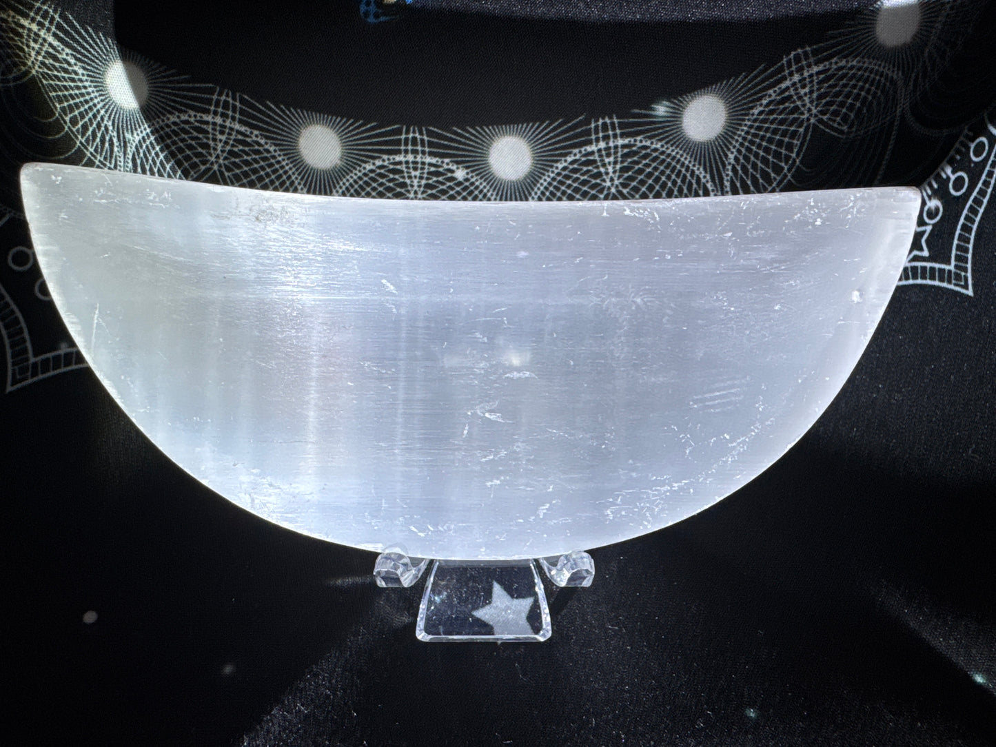 Selenite crescent bowl on a clear stand with a star pattern, resting on a black patterned cloth.