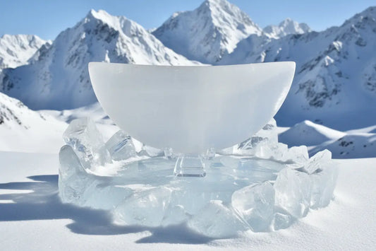 Selenite crescent bowl on a clear stand resting on a bed of ice with snowy mountains in the distance