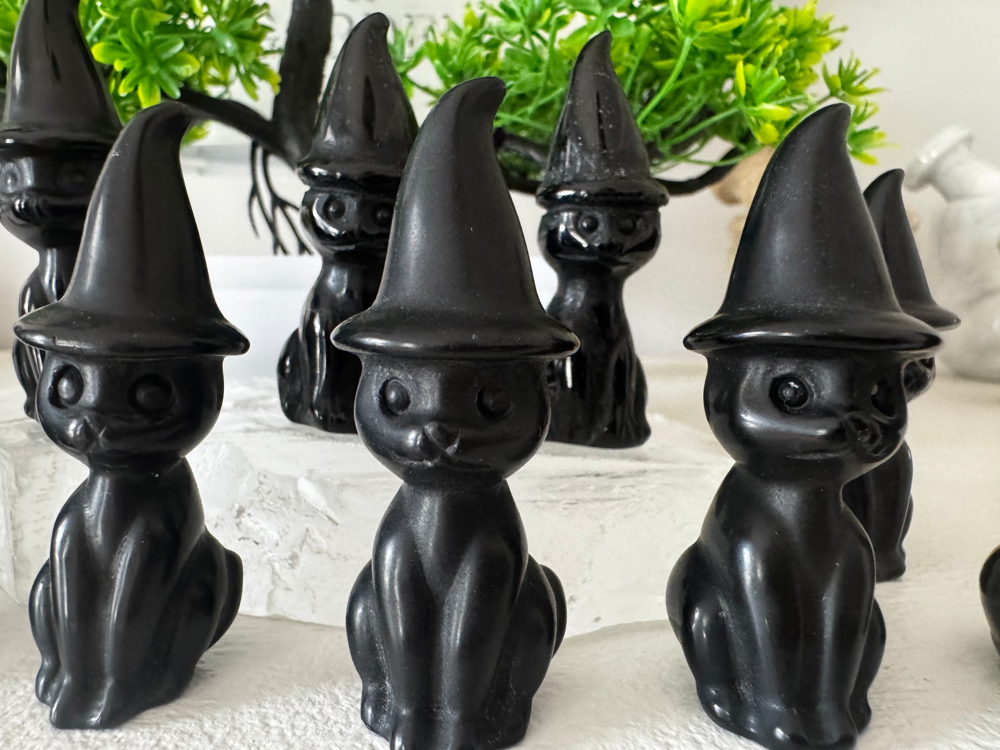 A group of glossy black, carved cats wearing pointed witch hats sitting on a white surface with green plants in the background.