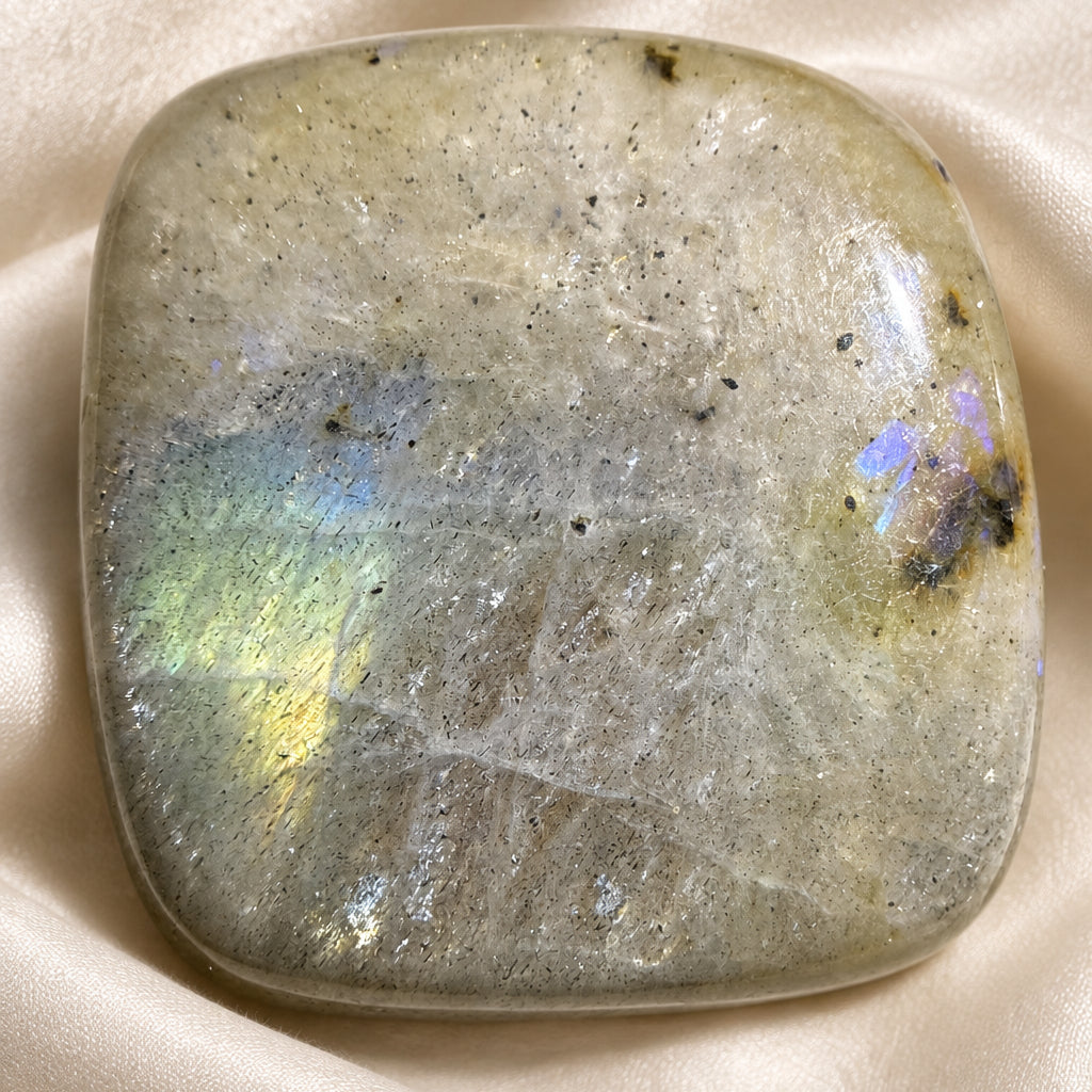 A polished natural labradorite stone cabochon with a rounded square shape, displaying a grayish-green base color and a shimmering, iridescent rainbow sheen on the left and right.