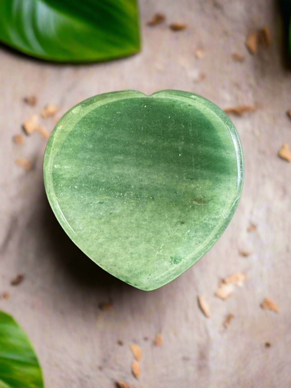 Heart-shaped green gemstone on a textured surface with scattered debris and leaf edges.
