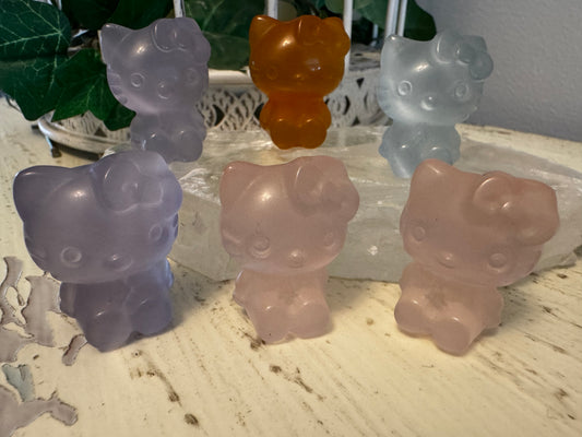 Six Hello Kitty shaped figurines made of translucent stones in varying shades of purple, pink, orange, and blue. They are arranged on a white surface with a piece of white stone between the front row and the back row. Some green leaves are visible in the background.