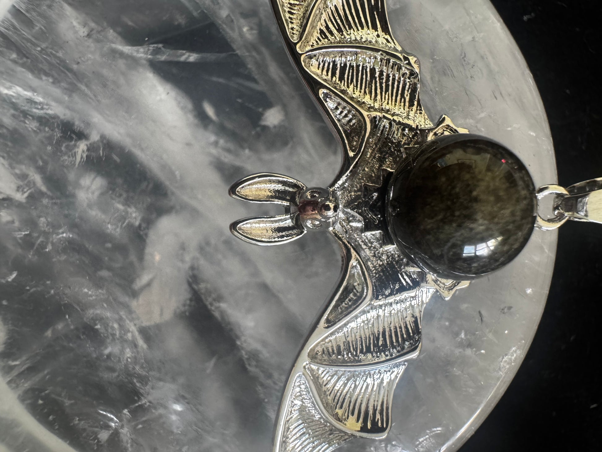 Close-up of a silver bat pendant with a dark, iridescent gemstone orb, resting on a textured, grey surface.