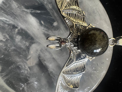 Close-up of a silver bat pendant with a dark, iridescent gemstone orb, resting on a textured, grey surface.