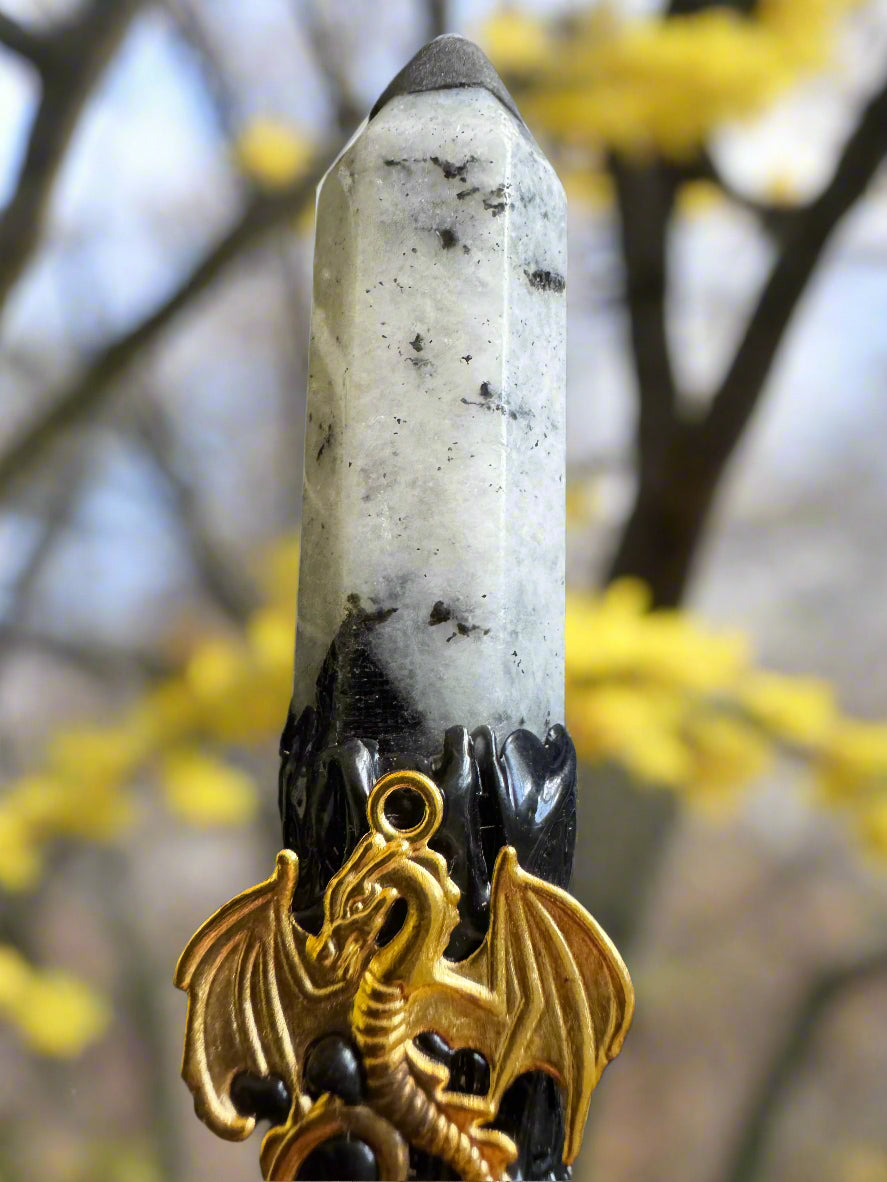 Close-up of a white crystal point with black accents, a black stand, and a gold-colored dragon charm, set against a blurry background of yellow flowers and tree branches.
