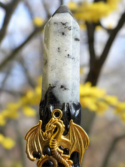 Close-up of a white crystal point with black accents, a black stand, and a gold-colored dragon charm, set against a blurry background of yellow flowers and tree branches.