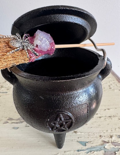 Close-up of a black, three-legged cauldron with a lid, adorned with a small broom featuring a silver spider and a purple crystal.