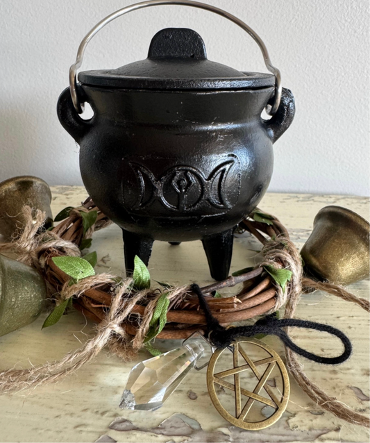 Triple moon pentacle black cast iron cauldron with lid, three legs, and metal handle, surrounded by rustic altar decor, crystal point, and gold pentagram ornament—perfect for witchcraft rituals, spellwork, and Wiccan altar supplies