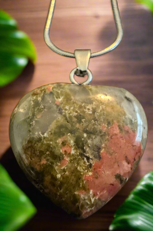 Heart-shaped pendant necklace made of a green and pink stone, hanging in front of a wooden background with green leaves.