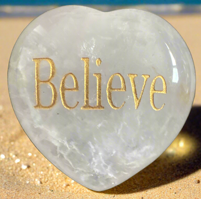 Heart-shaped crystal with "Believe" engraved in gold, on a sandy beach.
