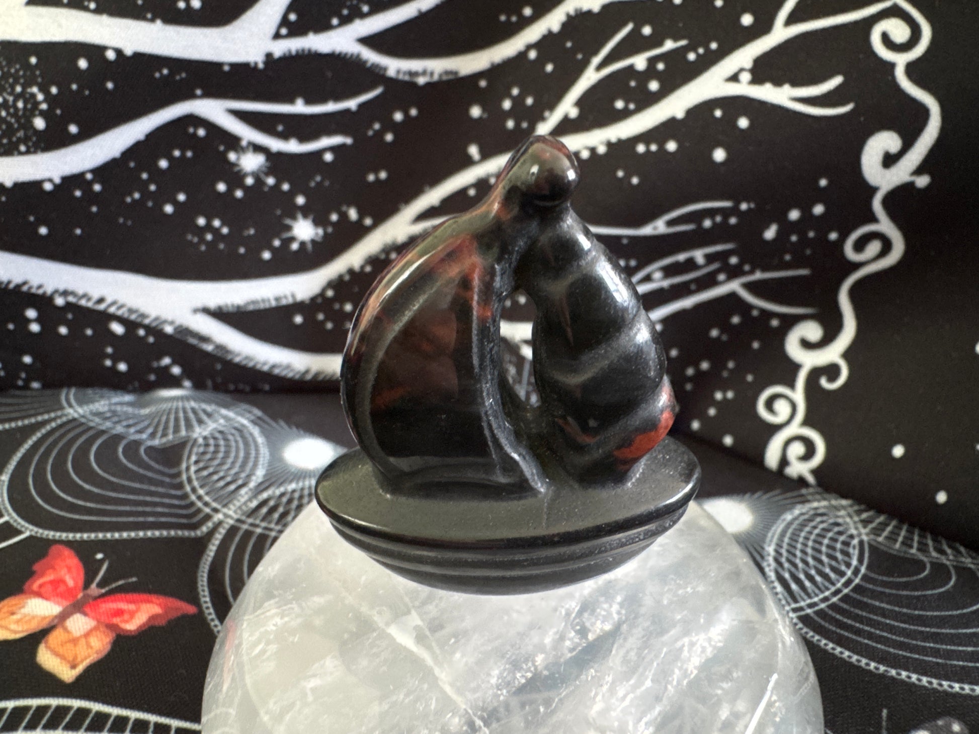 Black, translucent cat figurine on a round base atop a clear quartz crystal. Background is black with white patterns.