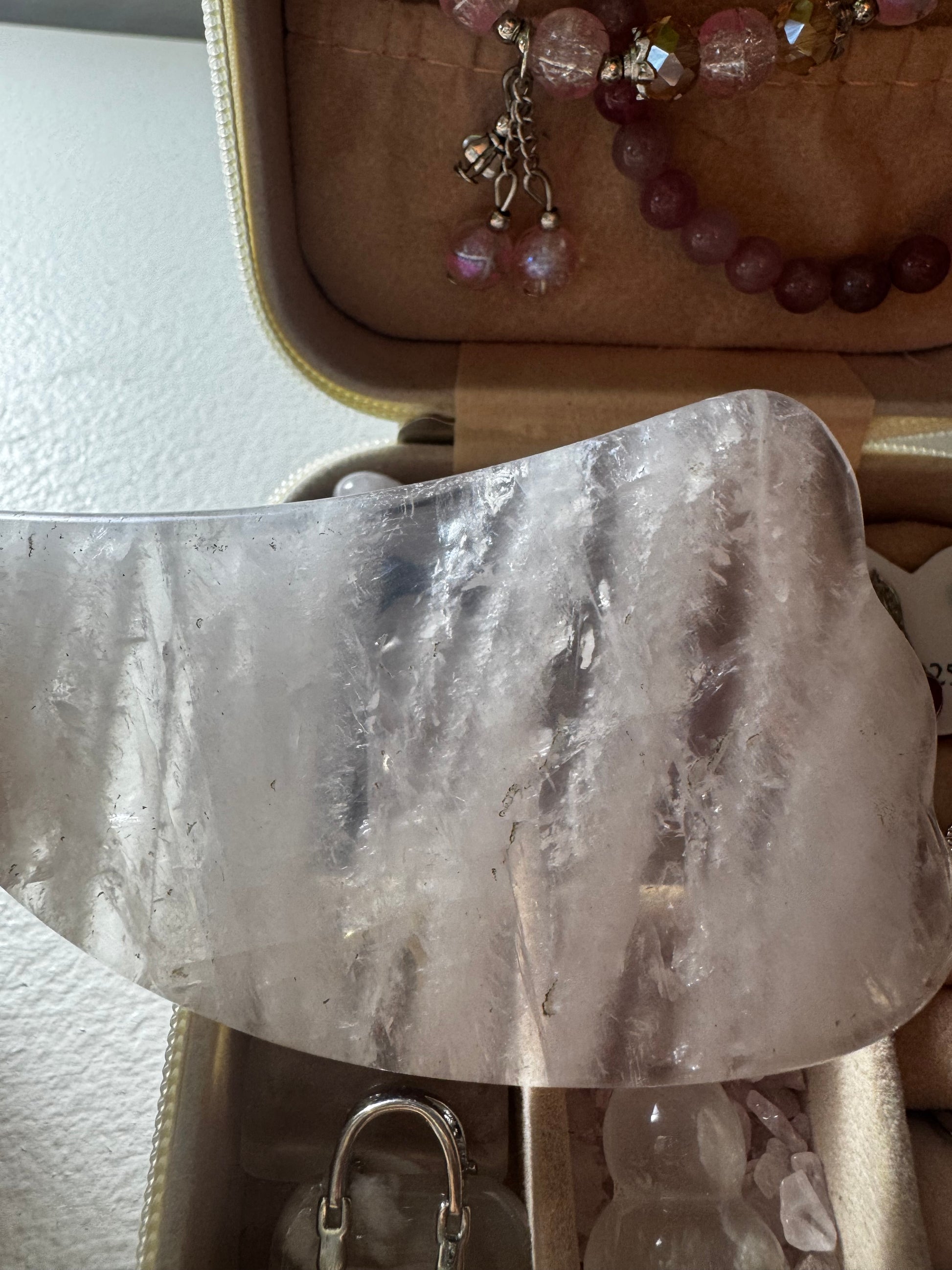 Close-up of a rose quartz gua sha tool in a jewelry box next to a pink beaded bracelet.