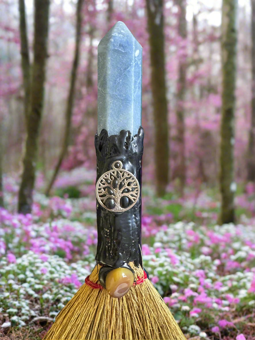 A handmade besom with a blue crystal, a tree charm, and a polished stone in a flower field.