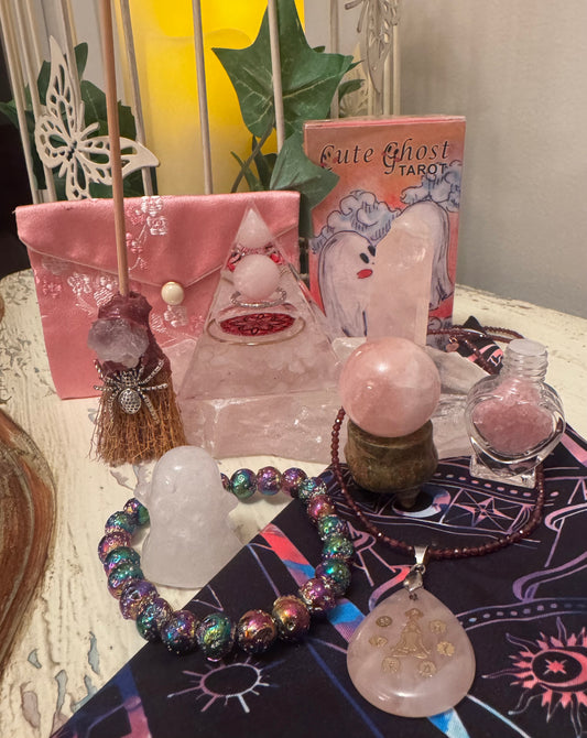 A collection of pink and iridescent spiritual objects, including crystals, a deck of "Cute Ghost Tarot" cards, a heart-shaped bottle with pink contents, and jewelry, arranged on a dark cloth with astral-themed patterns.