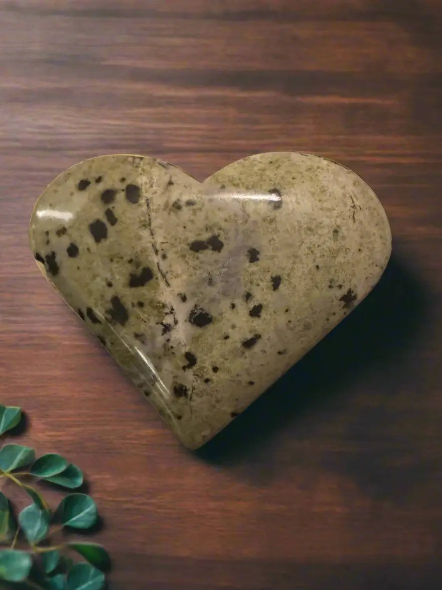Heart-shaped, mottled green stone on wooden surface with leafy green plant.
