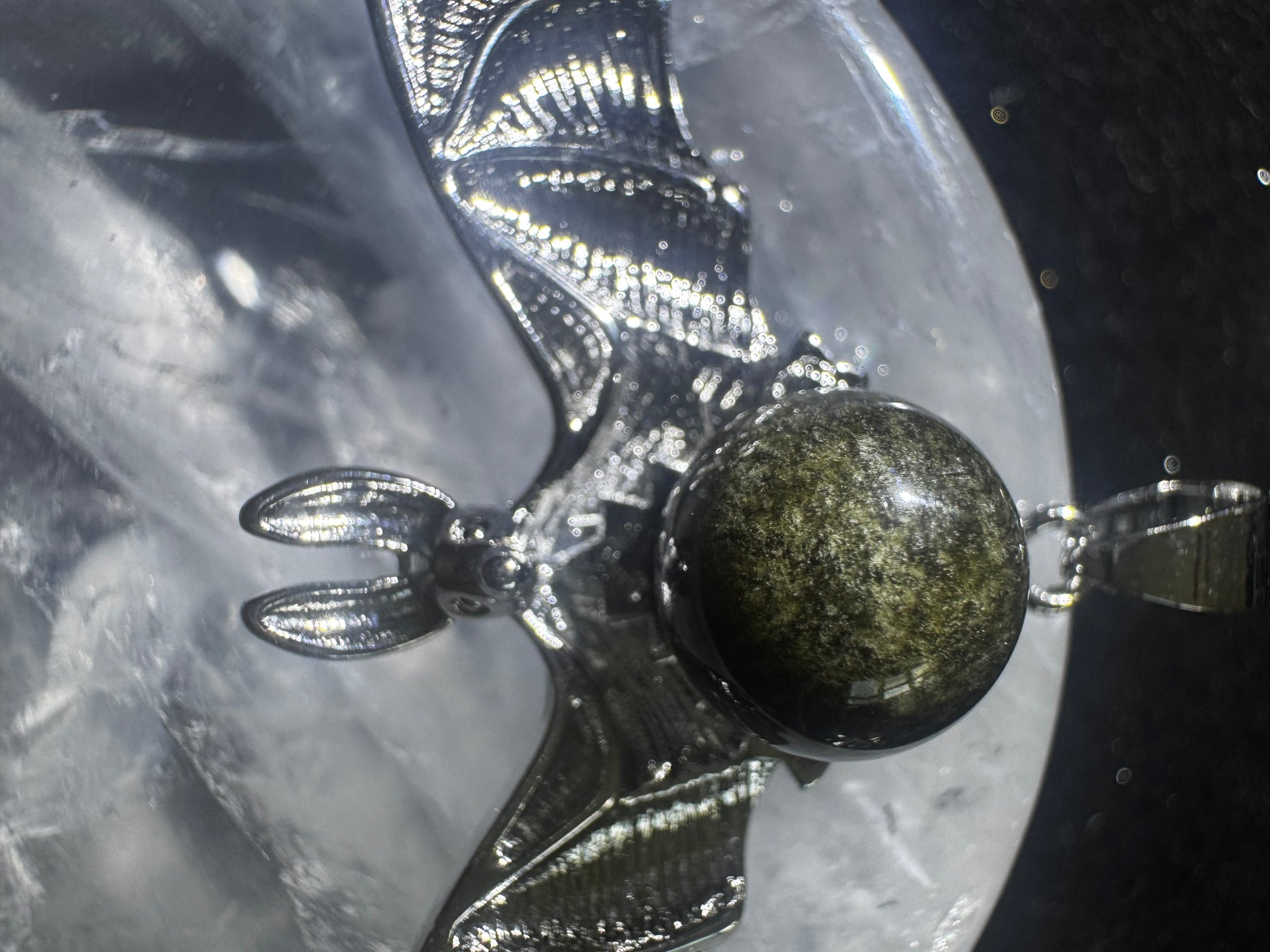 Close-up of a silver bat pendant featuring a green and brown speckled stone set against a quartz background.