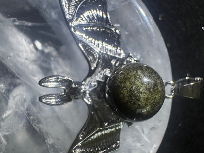 Close-up of a silver bat pendant featuring a green and brown speckled stone set against a quartz background.