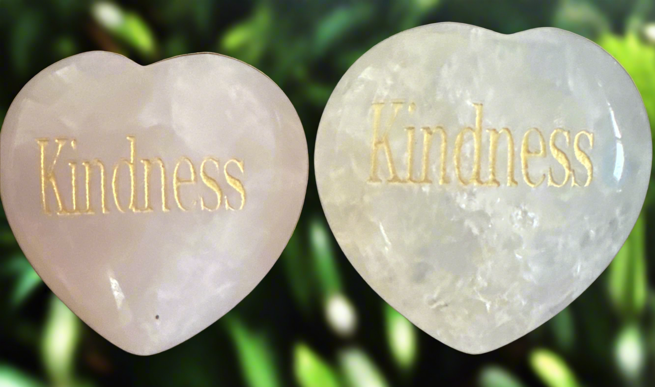 Two heart-shaped stones with "Kindness" engraved in gold, against a blurred green leafy background.