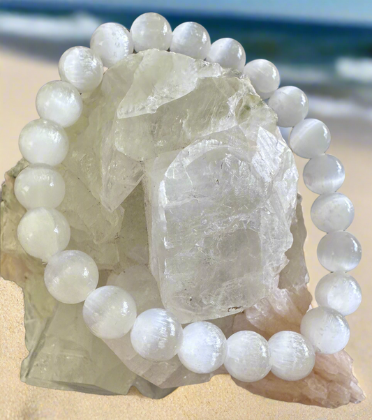 A white crystal bead bracelet is draped over a large, clear quartz crystal cluster on a beach. The beach backdrop features tan sand, blue water, and sky.