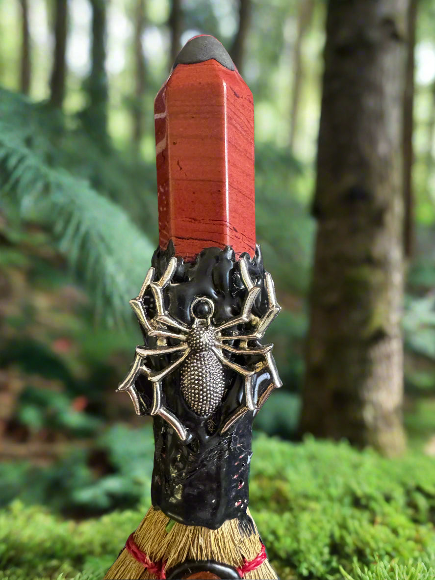 Close-up of a small decorative broom with a red stone handle, featuring a silver spider embellishment and black resin detailing, set in a natural forest environment.
