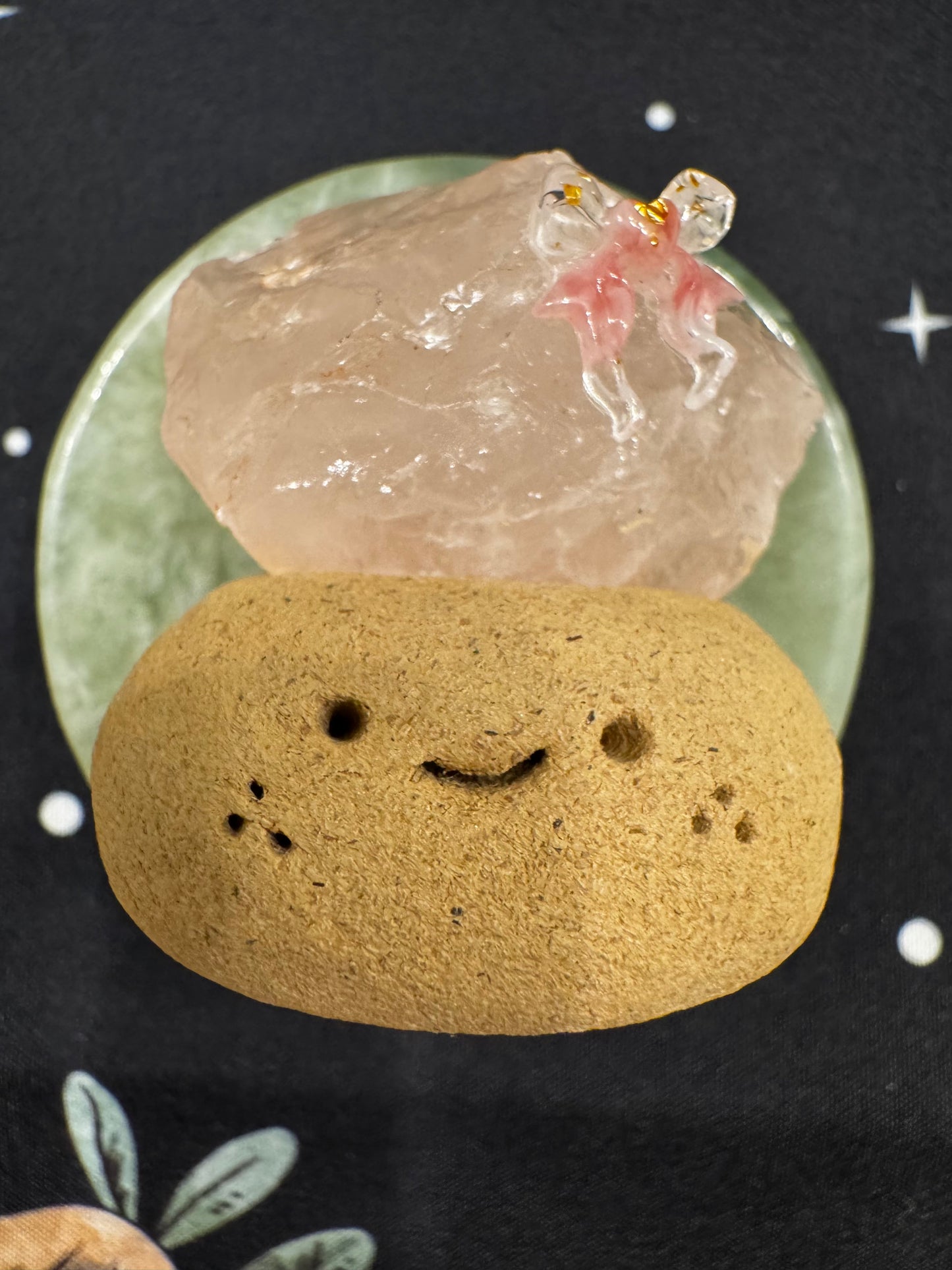 A smiling tan stone with holes sits beneath a pink crystal with a pink and gold figure on a green dish.
