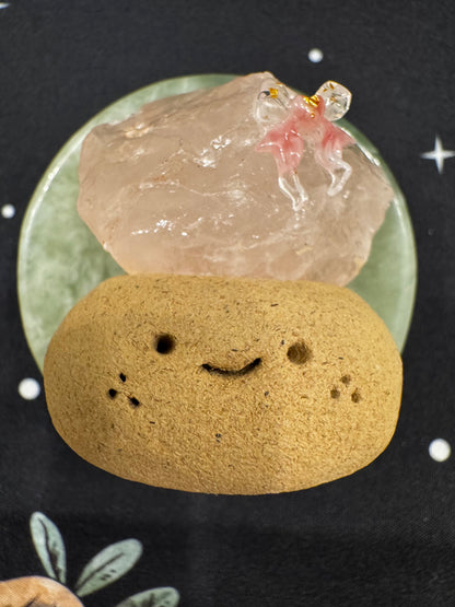 A smiling tan stone with holes sits beneath a pink crystal with a pink and gold figure on a green dish.