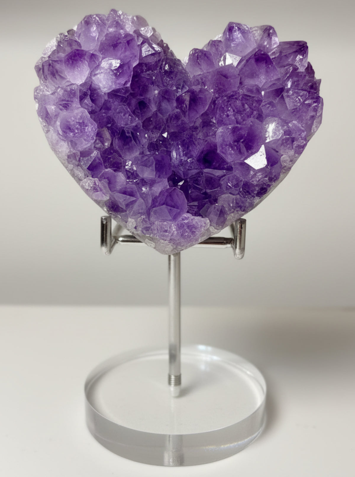 A heart-shaped amethyst crystal cluster on a clear acrylic stand, displayed on a light-colored wooden surface against a neutral background.