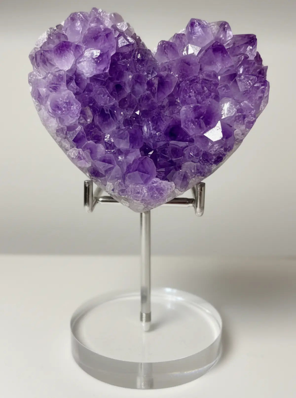 A heart-shaped amethyst crystal cluster on a clear acrylic stand, displayed on a light-colored wooden surface against a neutral background.