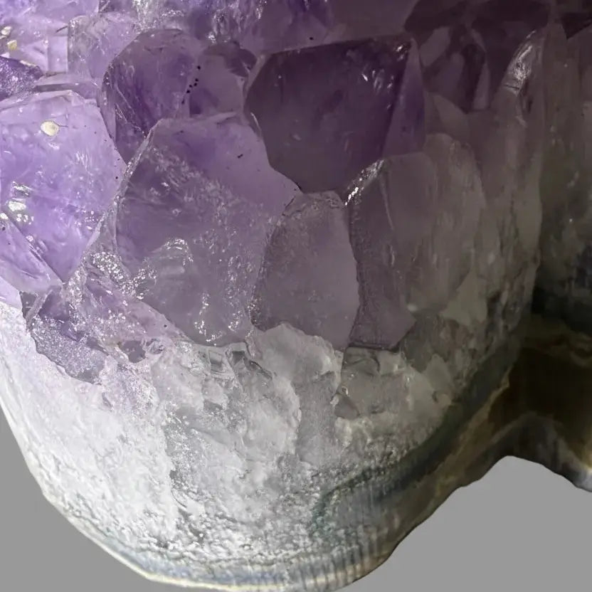 Close-up of an amethyst crystal cluster with translucent purple crystals and a white textured base, photographed against a plain gray background.