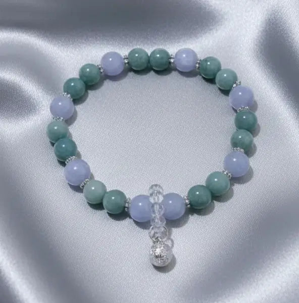 A beaded stretch bracelet featuring angelite light blue and aventurine round beads, accented with small silver spacer beads and a dangling protection bell.   