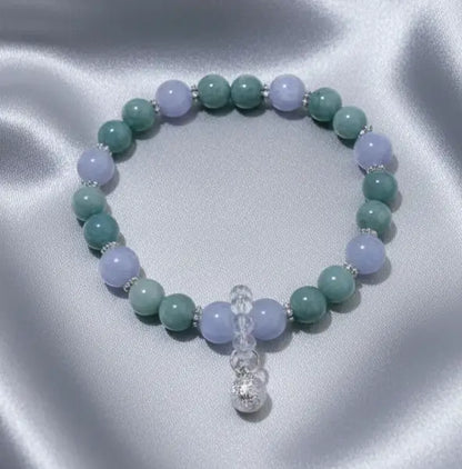 A beaded stretch bracelet featuring angelite light blue and aventurine round beads, accented with small silver spacer beads and a dangling protection bell.   