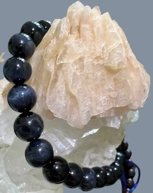 A beaded bracelet made of glossy dark blue to black round beads, draped over a pale pink crystal cluster against a plain gray background.