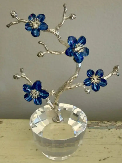 A decorative crystal tree  with silver-toned branches, four blue faceted flower blossoms, and a clear faceted glass base, shown against a neutral background.