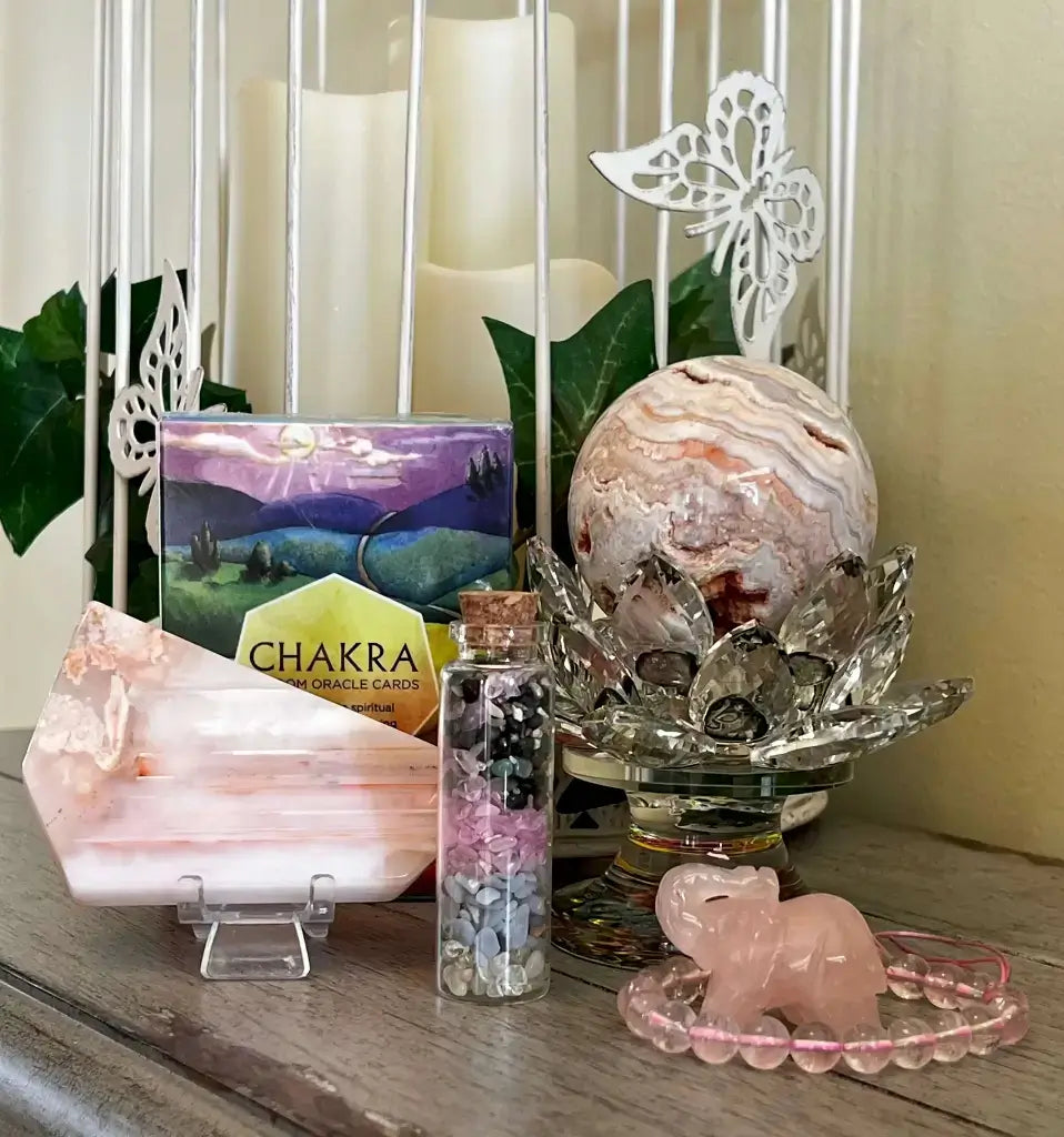 A styled arrangement of crystals and spa accessories on a wooden surface, including a pink agate crystal cluster, a pink elephant-shaped bead holder, a glass vial with colorful stones, a large agate sphere on a lotus stand, and a chakra card deck box.