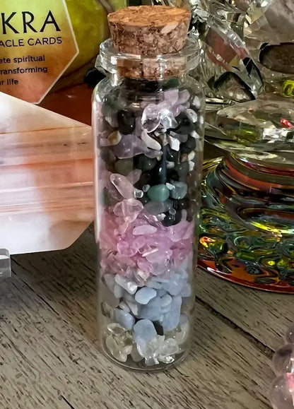 A small clear glass bottle with a cork stopper, filled with layered crystals and stones in pink, white, black, and pale blue, placed on a wooden surface.