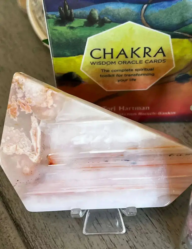 A triangular polished agate crystal display stand with natural peach and white banding, shown on a small clear acrylic easel in front of a Chakra Wisdom Oracle Cards box on a wooden surface.