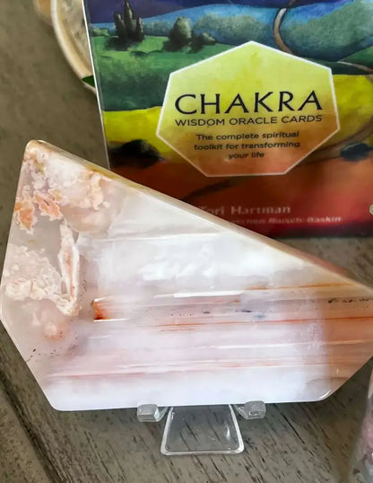 A triangular polished agate crystal display stand with natural peach and white banding, shown on a small clear acrylic easel in front of a Chakra Wisdom Oracle Cards box on a wooden surface.
