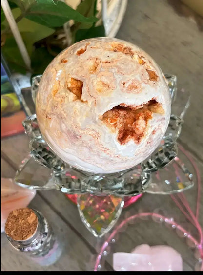 A polished crystal sphere with swirling white, cream, and copper-orange marbling, displayed on a clear faceted glass stand over a decorative base.