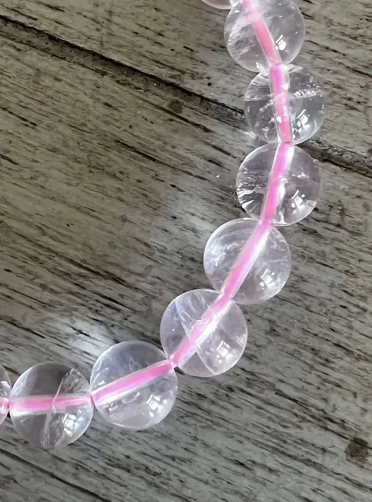 Clear round gemstone beads threaded on a bright pink cord, laid on a weathered gray wooden surface.