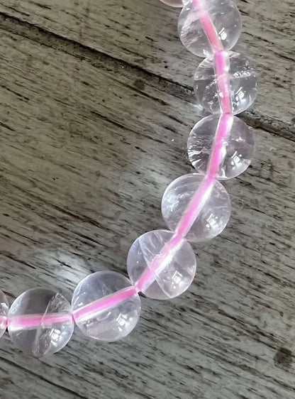 Clear round gemstone beads threaded on a bright pink cord, laid on a weathered gray wooden surface.