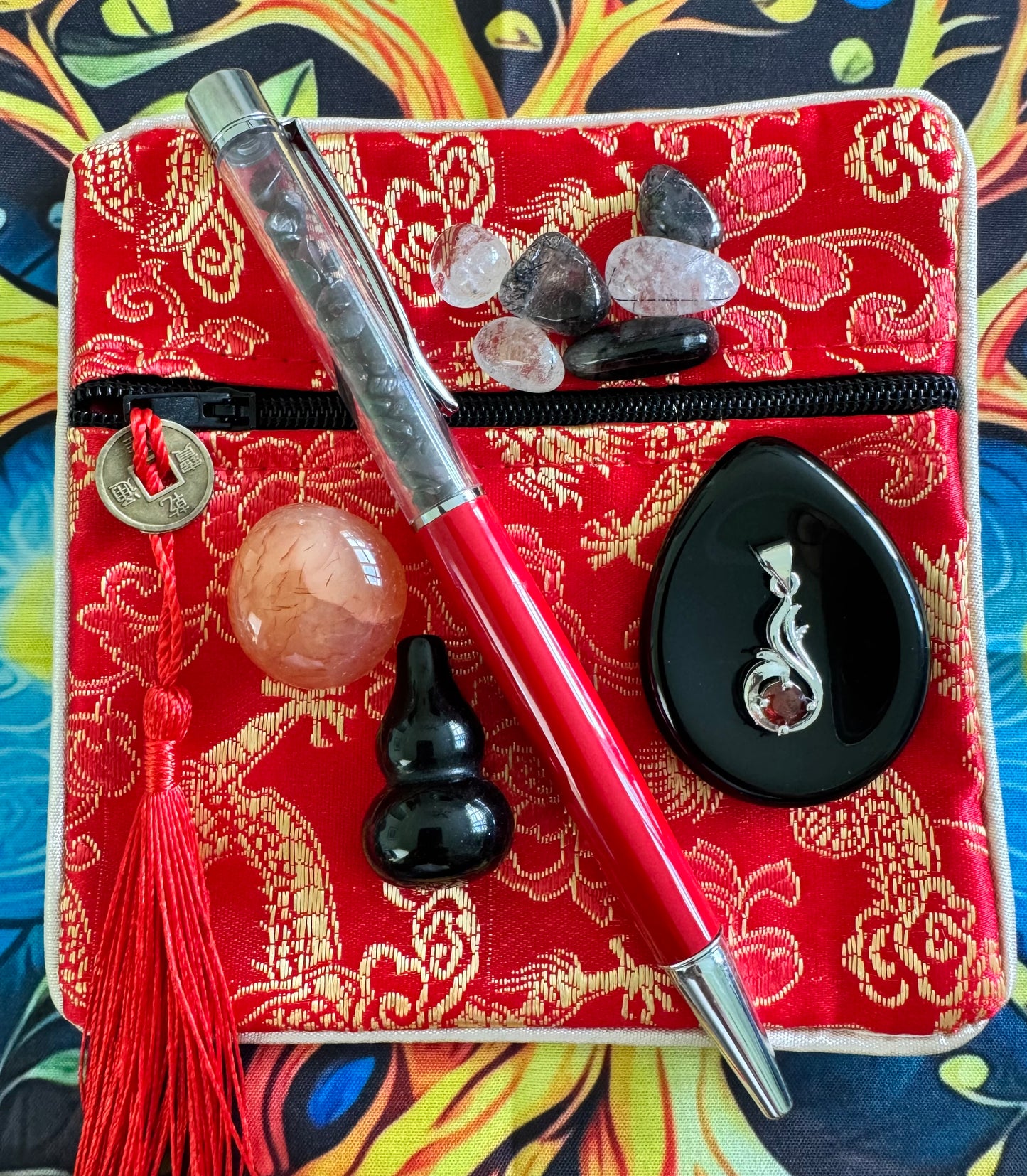 A red zippered bag with a gold floral pattern, accented with a coin tassel, crystals, a red pen filled with crystals, a peach-colored sphere, a black gourd, and a black pendant with a garnet.