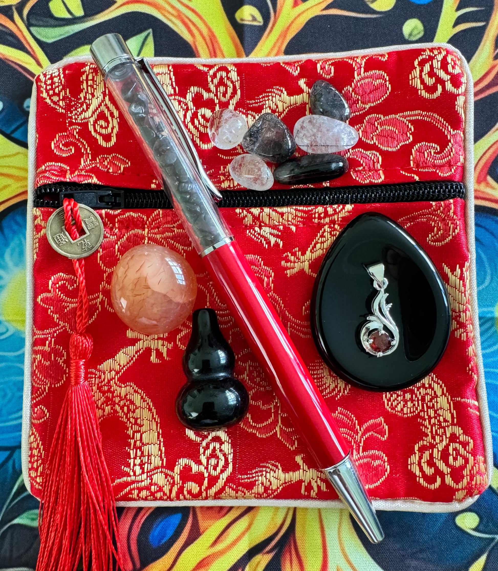 A red zippered bag with a gold floral pattern, accented with a coin tassel, crystals, a red pen filled with crystals, a peach-colored sphere, a black gourd, and a black pendant with a garnet.