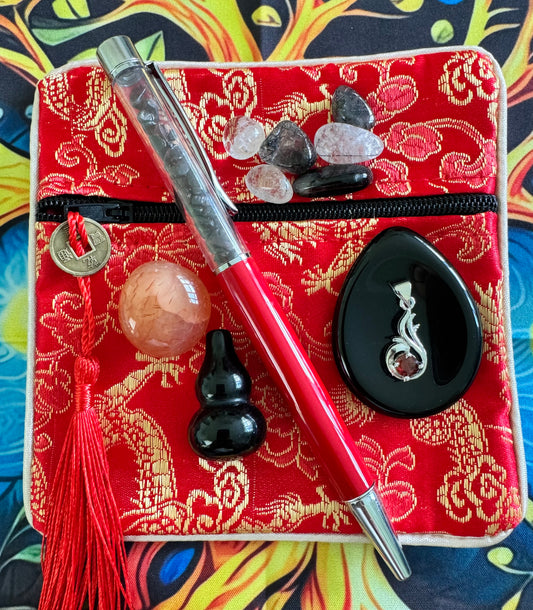 A red zippered bag with a gold floral pattern, accented with a coin tassel, crystals, a red pen filled with crystals, a peach-colored sphere, a black gourd, and a black pendant with a garnet.