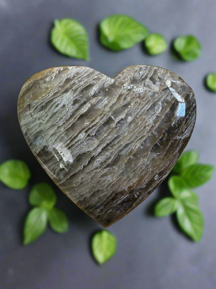 Heart-shaped crystal, striated pattern, surrounded by green leaves, gray background.
