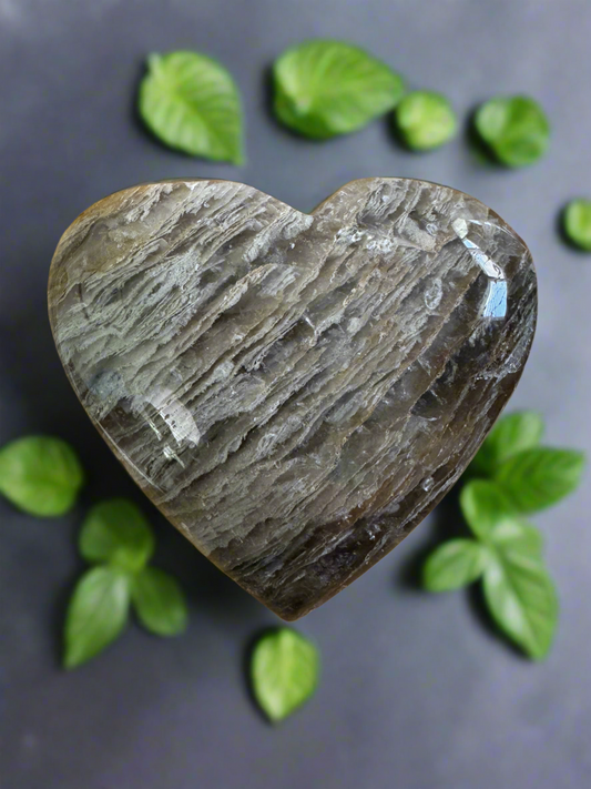 Heart-shaped crystal, striated pattern, surrounded by green leaves, gray background.
