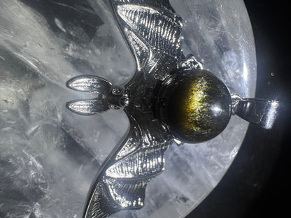 A silver bat pendant with a golden sheen rests on a clear crystal.