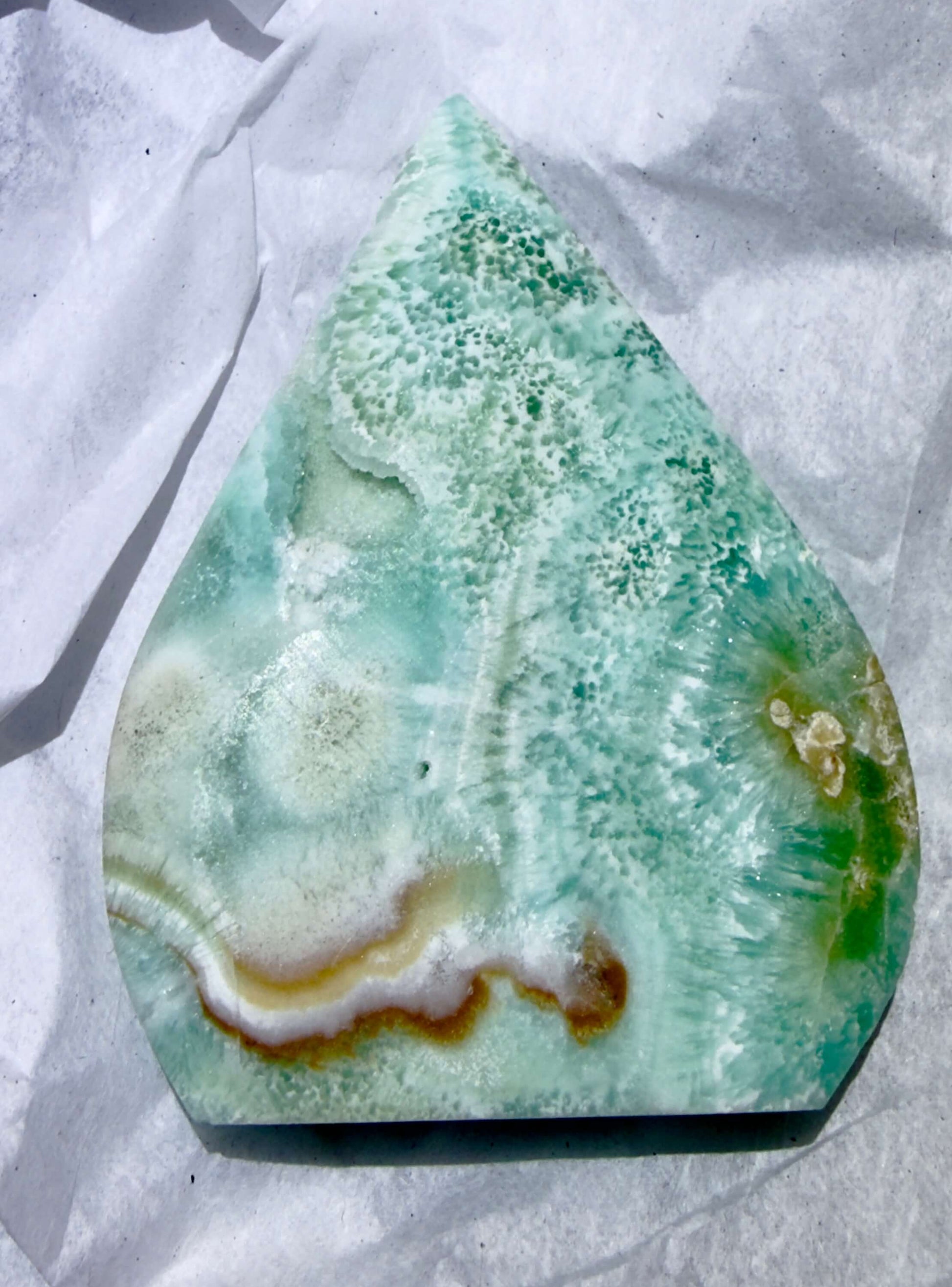 A polished teardrop-shaped caribbean calcite freeform stone with green, white, and brown banding, displayed on crumpled white paper.