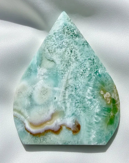 A polished teardrop-shaped caribbean calcite freeform stone with green, white, and brown banding, displayed on crumpled white paper.