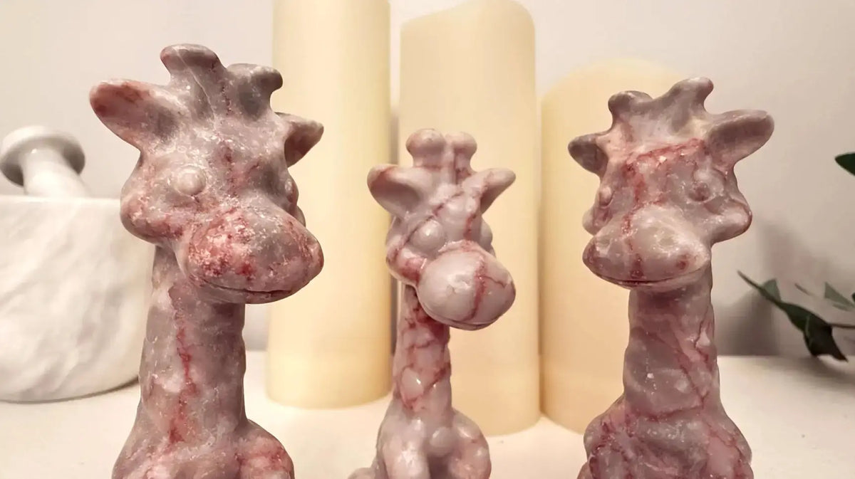 Three small giraffe figurines made of network, also know as,  red vein jasper arranged in front of cream-colored pillar candles on a tabletop.