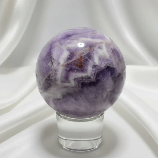 Purple, White, clear chevron amethyst carved sphere with iron inclusions on a glass stand with a white fabric in the background